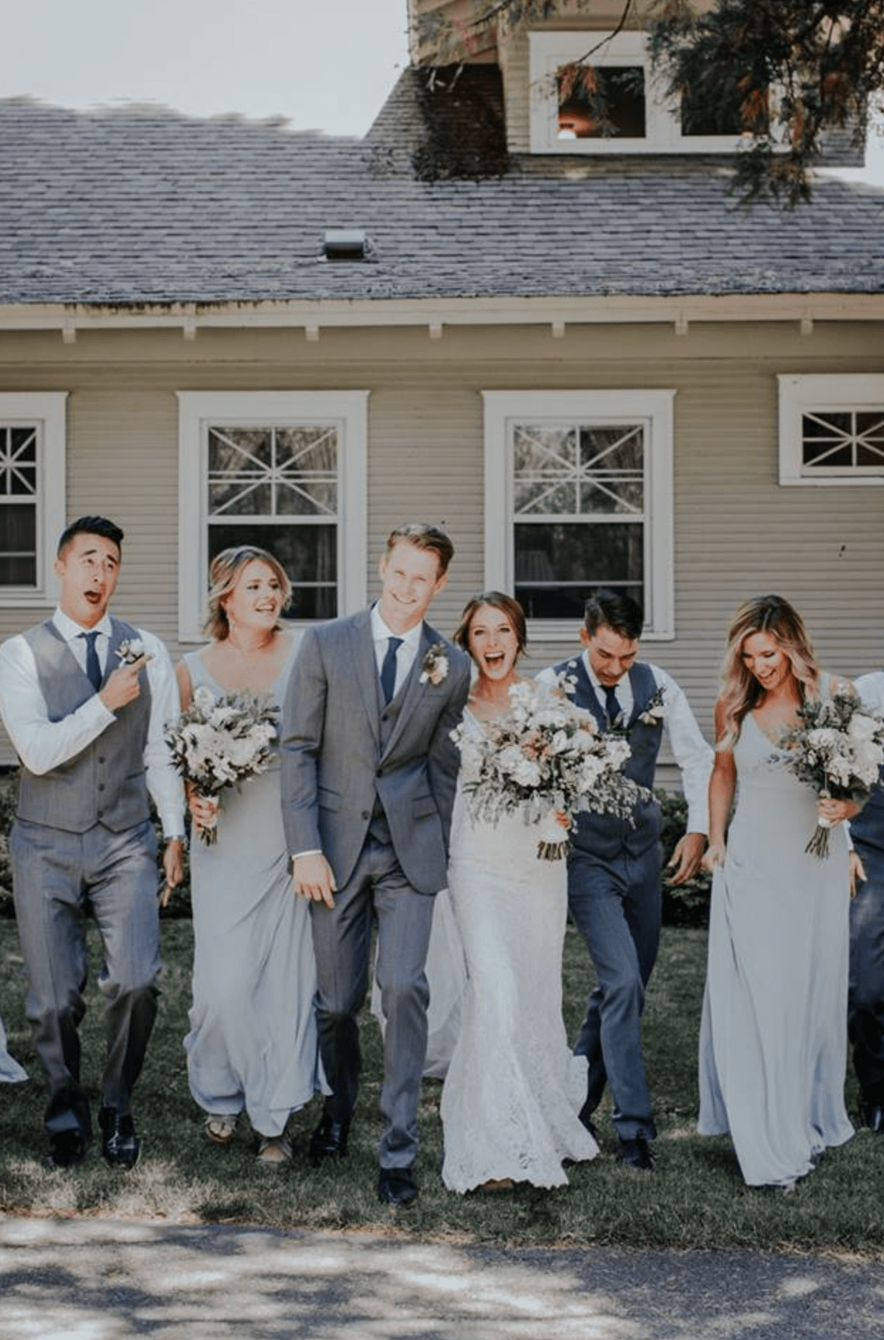 A joyous wedding party joyfully walks together outdoors, holding bouquets and smiling in front of a house.