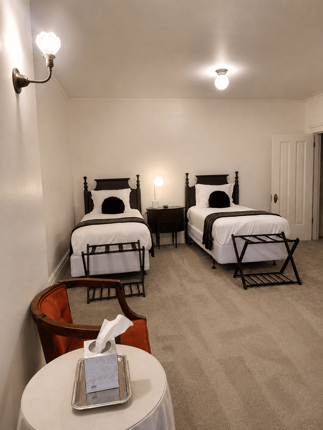A cozy bedroom featuring two twin beds with dark throw pillows, a nightstand, and a chair by a small table with tissues.