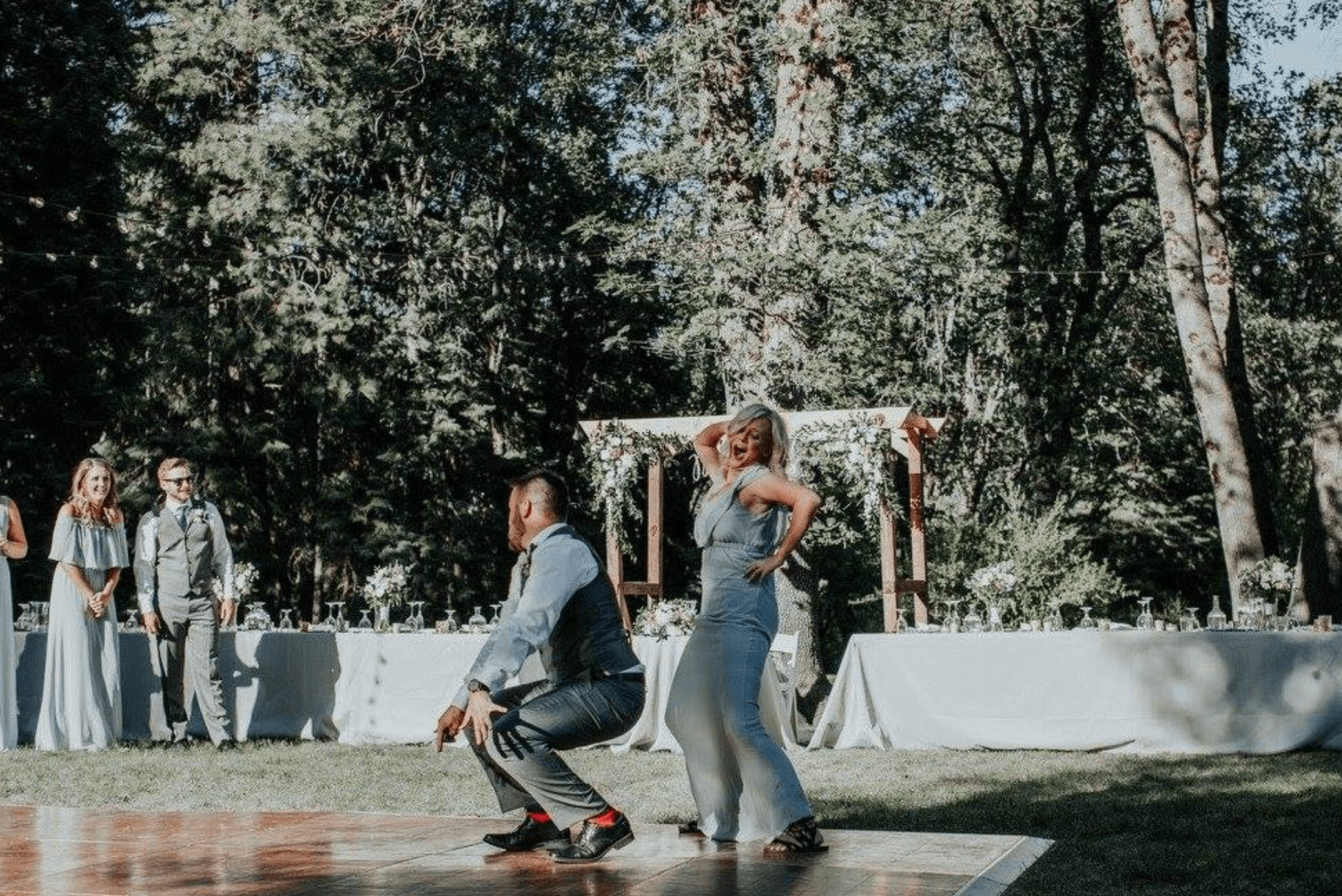 A man and woman dance joyfully at an outdoor wedding, surrounded by guests in formal attire.