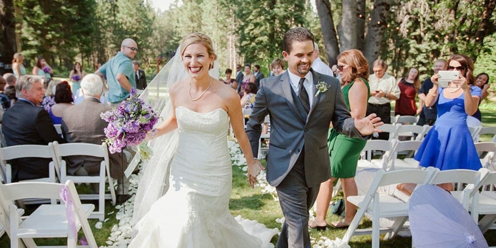 A smiling bride and groom walk hand in hand down an outdoor aisle, surrounded by guests celebrating their wedding.