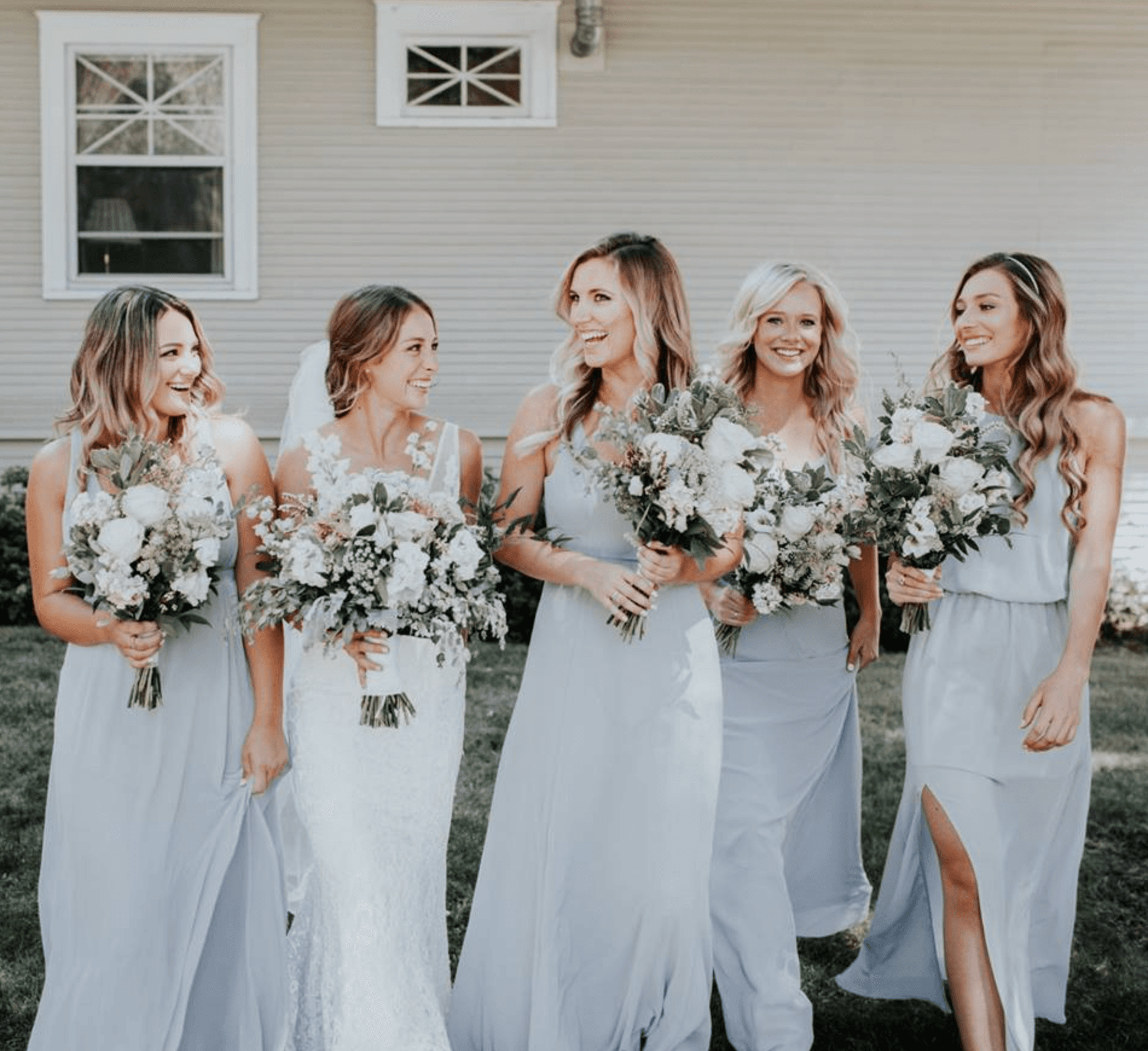 A bride and her bridesmaids in gray dresses hold bouquets while walking together outdoors.