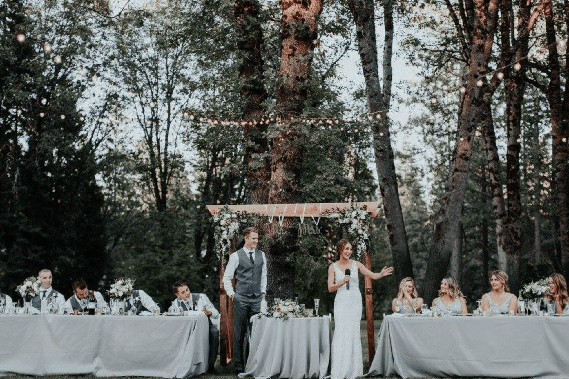 A bride delivers a toast at an outdoor wedding reception surrounded by guests at elegantly set tables.