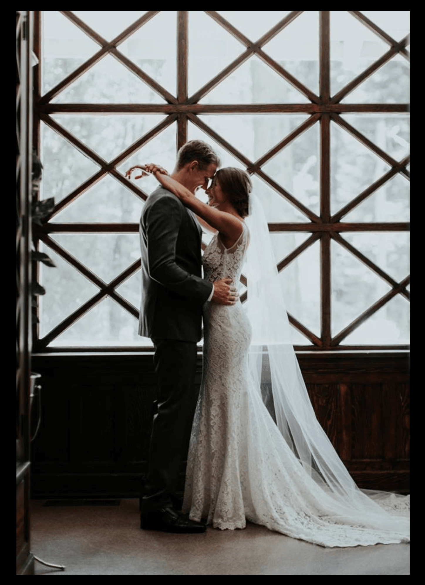 A bride and groom embrace in a romantic moment by a large, lattice window.