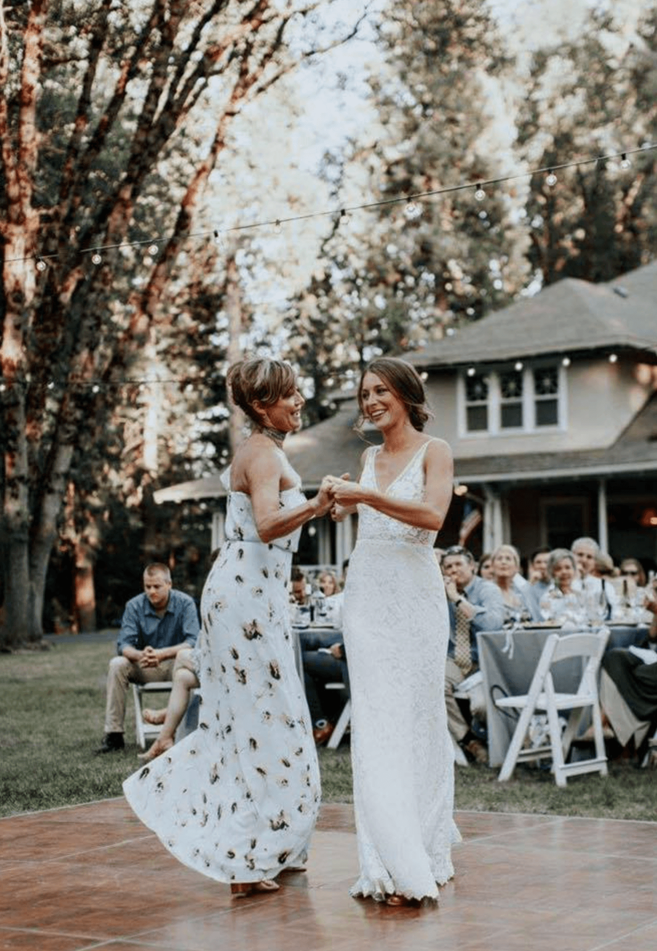 Two women dance joyfully on a wooden floor, surrounded by guests at an outdoor celebration.