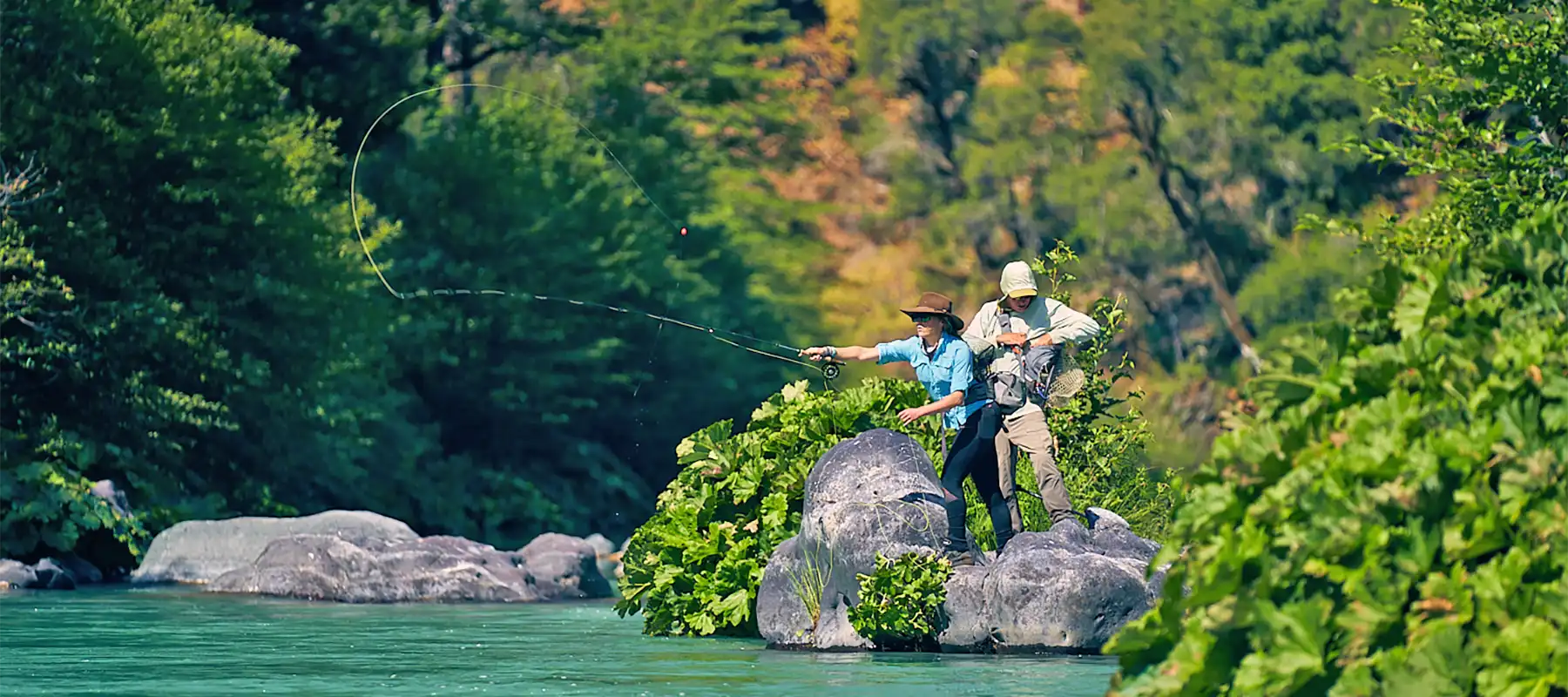 Two fishermen casting lines from a rock by a lush, green riverbank.
