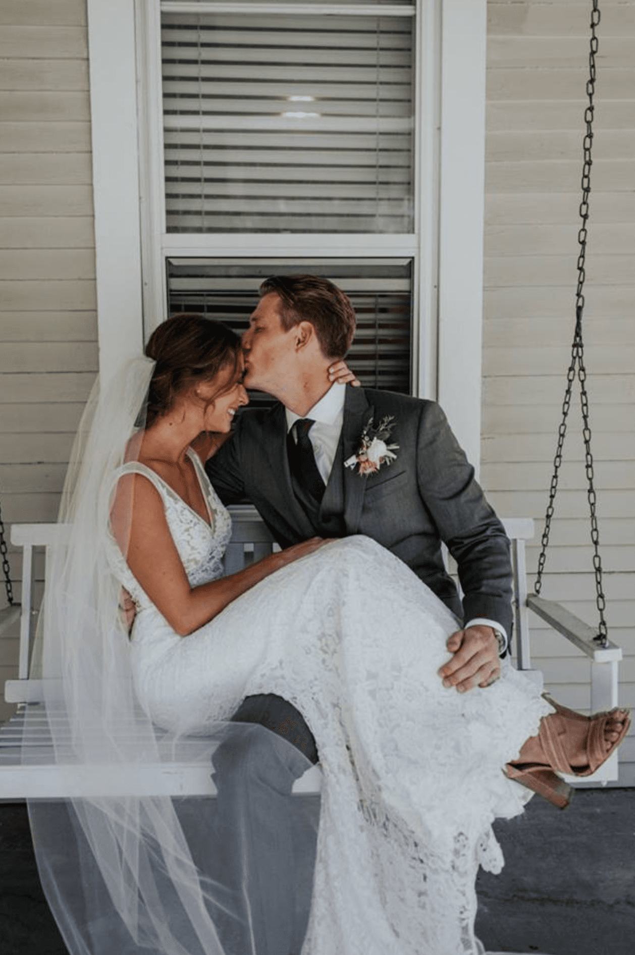A newlywed couple shares a tender kiss while sitting together on a porch swing.