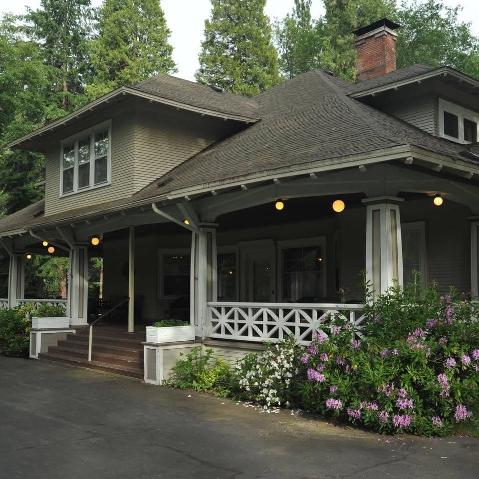 A two-story house with a large porch, surrounded by trees and flowering plants.