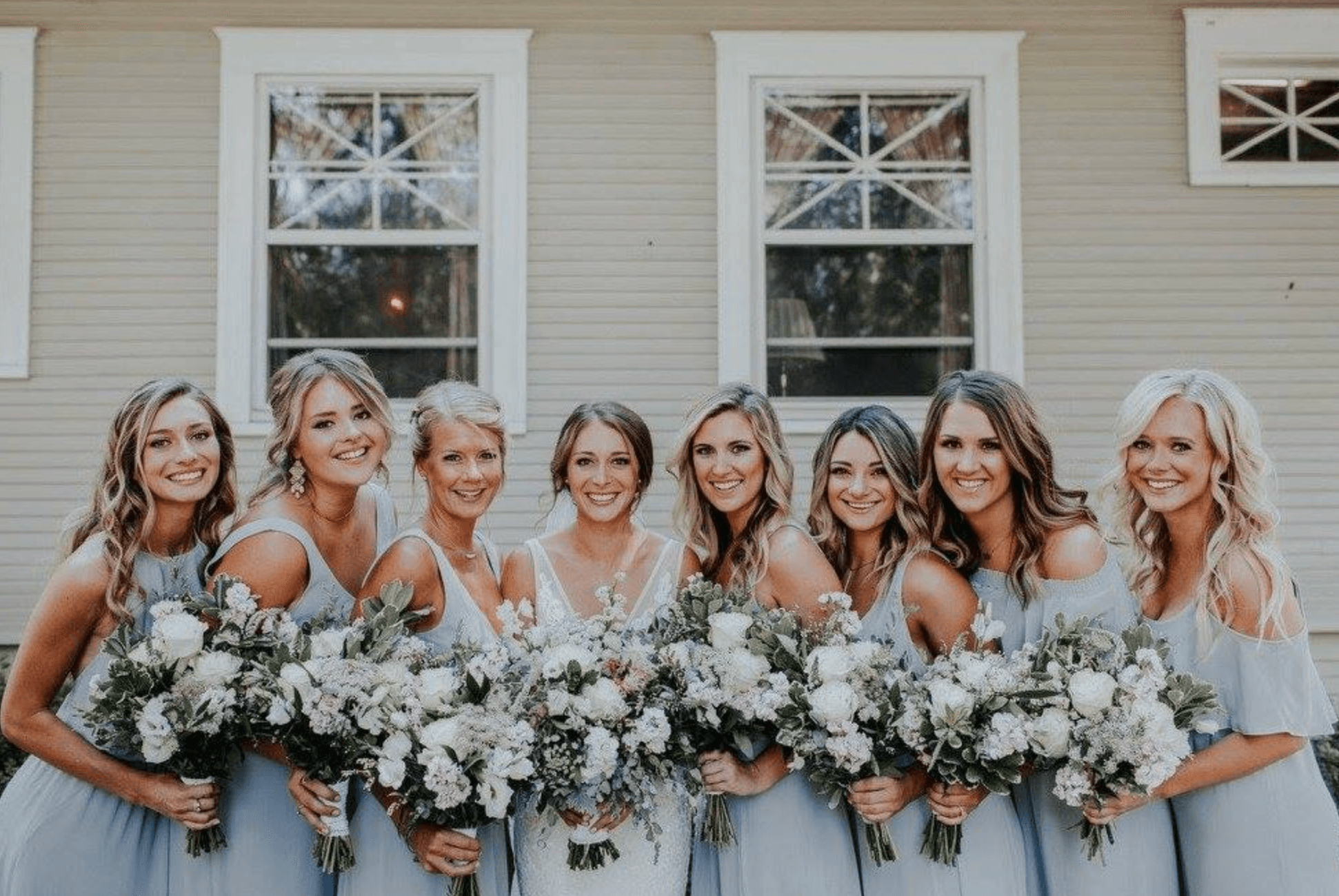 A group of eight women in light blue dresses holds bouquets while posing together in front of a house.