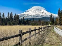 Snow-capped mountain rises above a grassy field and wooden fence under a blue sky.
