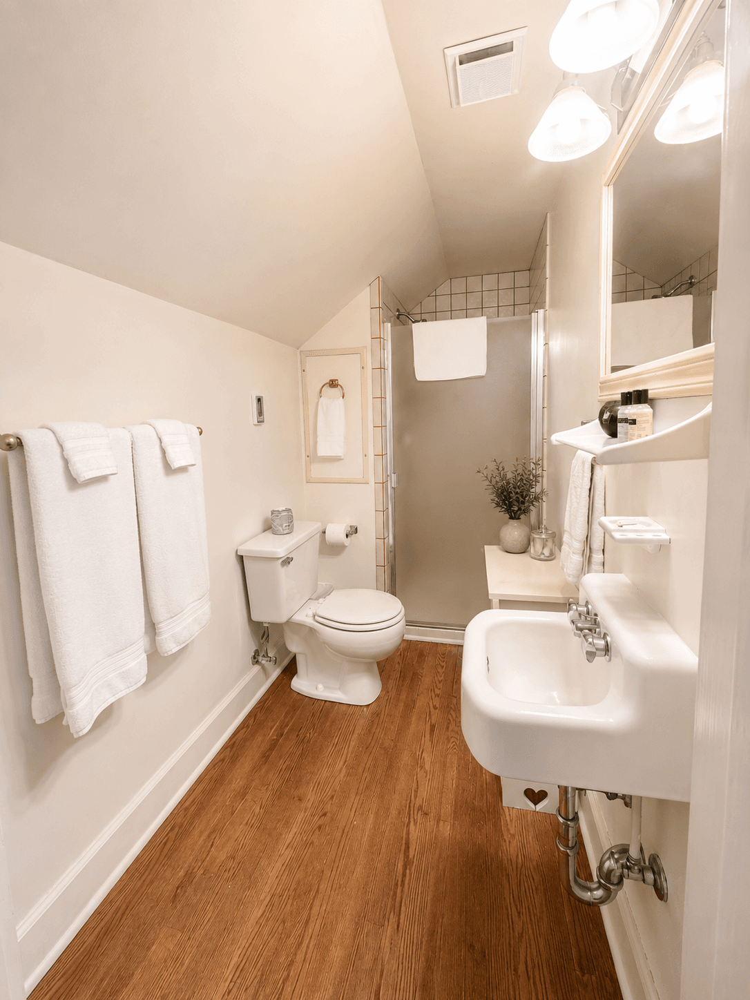 A compact bathroom featuring a toilet, a small sink, and a shower, with wooden flooring and white towels.